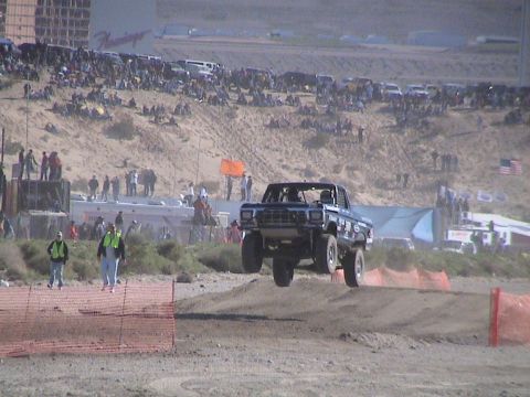 1990 Ford Bronco - black Ford pickup catches air over a sand jump on a dirt course as a large crowd watches at a desert off-road event.