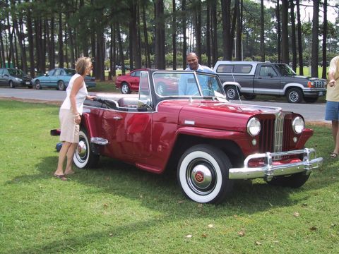 1968 Shelby GT500 - A red convertible Jeep is parked on grass with people nearby and other vehicles in the background.