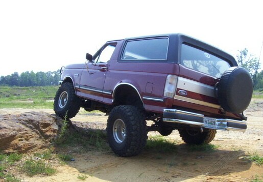 Lifted maroon 1990s Ford Bronco flexing over a dirt mound, rear three-quarter view with spare tire