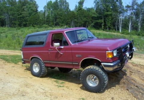 1989 Ford Bronco - This red Ford Bronco is shown from a 3/4 front angle, parked on a dirt incline with a wooded area behind it.