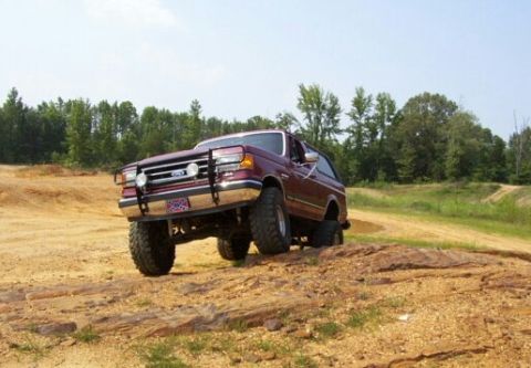 1989 Ford Bronco - A maroon Ford Bronco navigates a dirt terrain with a clear sky and trees in the background.