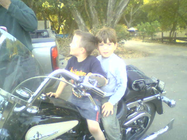Motorcycle with two young boys sitting on it, one in a dark t-shirt, the other in a light blue long-sleeved shirt