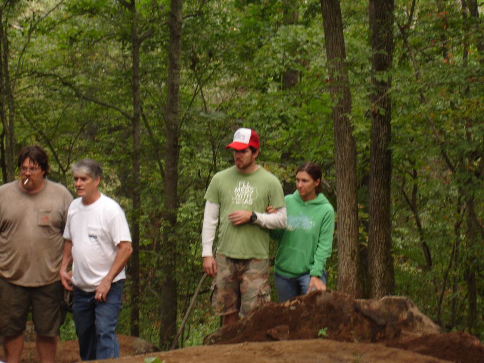 Four people stand in a wooded area with dirt mounds in the foreground