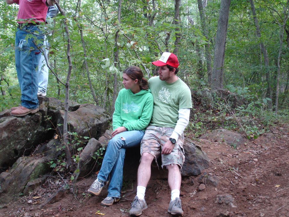Two people sit on rocks in a wooded area, with another person standing behind them