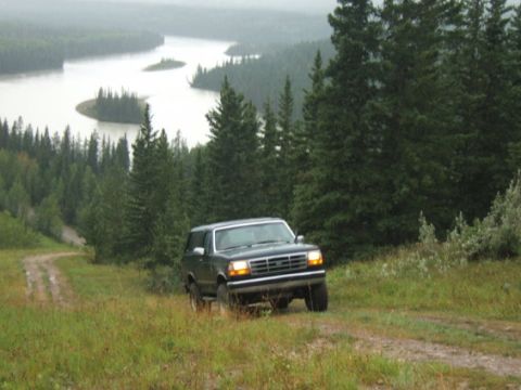 1995 Ford Bronco - A Ford Bronco navigates a dirt road with a scenic lake and forested hills in the background.