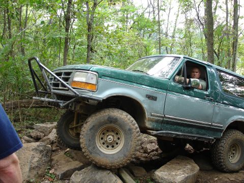1996 Ford Bronco - A green Ford Bronco is shown off-roading over large rocks in a wooded area.