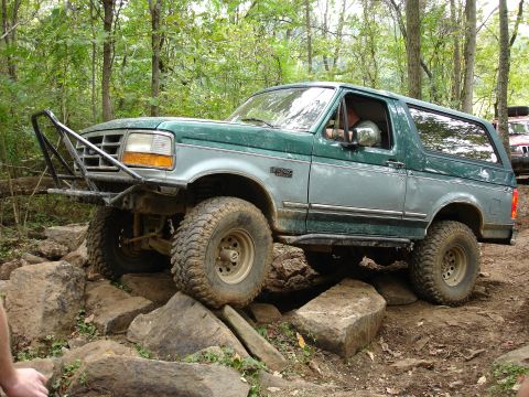 1996 Ford Bronco - 1996 Ford Bronco tackling an off-road trail with large rocks and trees in the background.