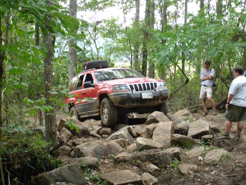1996 Ford Bronco - A red Jeep Grand Cherokee is shown tackling a challenging rocky terrain on an off-road trail.