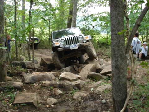 1996 Ford Bronco - A Jeep Wrangler tackles a challenging rocky path in a wooded area, with spectators watching.