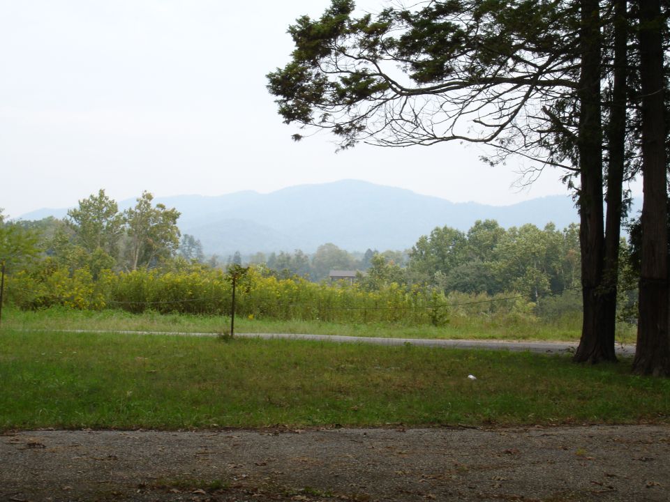 Misty mountains and trees overlook a grassy field with a distant house