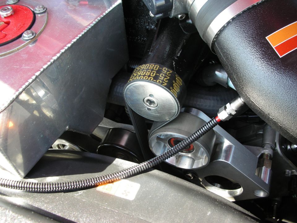 Engine bay close-up showing a black belt with yellow text, aluminum pulleys, and a red-capped fluid reservoir