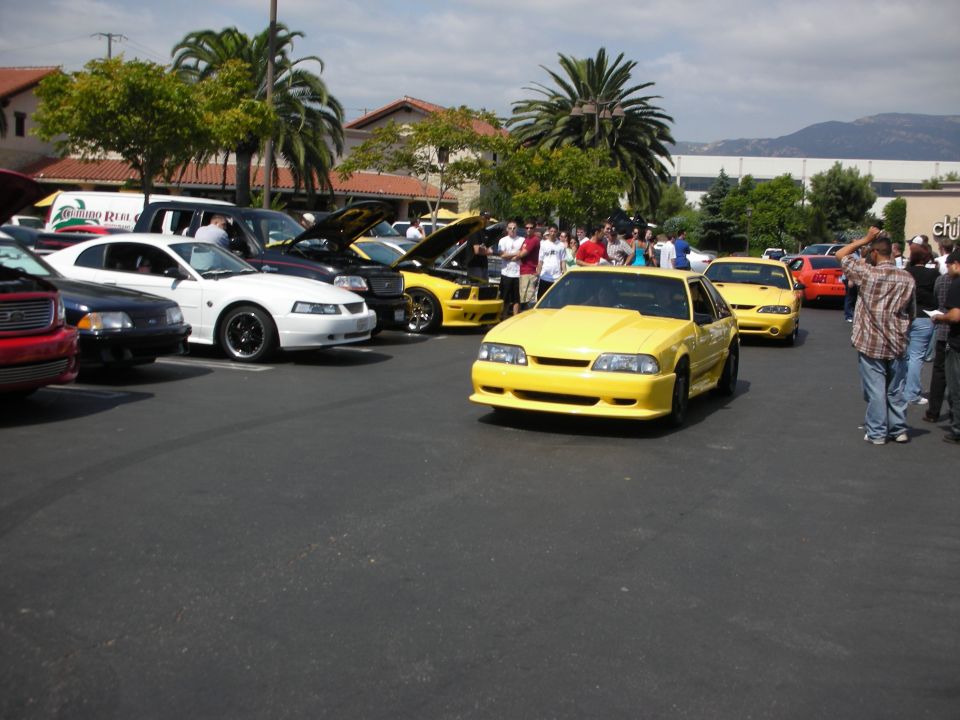 Yellow Ford Mustang with a hood scoop driving in a parking lot with other cars and people