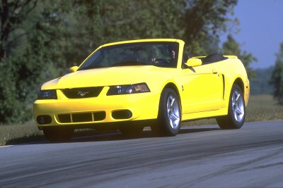 2003 Ford Mustang - A bright yellow 2003 Ford Mustang convertible is captured in a three-quarter front view on a paved road.