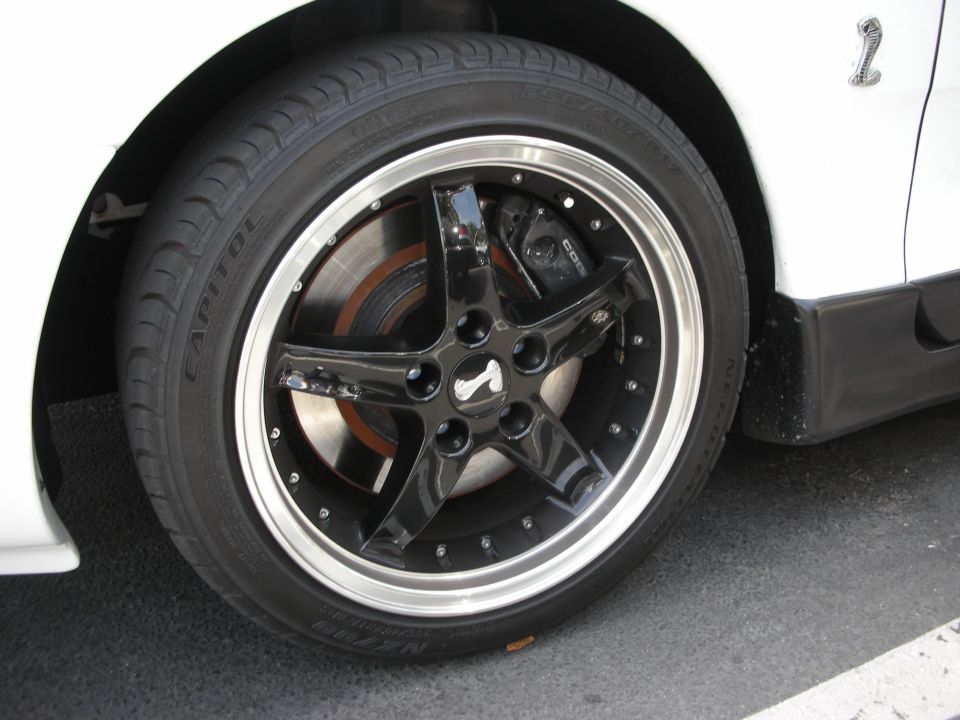Close-up of a black five-spoke wheel with a cobra emblem and performance brakes on a white car