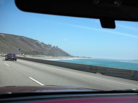 1992 Ford Crown Victoria - A dark sedan travels down a highway alongside a scenic coastline with the ocean on the right.