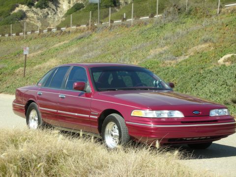 1992 Ford Crown Victoria - A maroon 1992 Ford Crown Victoria sedan is parked on a dirt road with a grassy hillside in the background.
