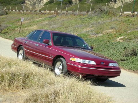 1992 Ford Crown Victoria - A red 1992 Ford Crown Victoria sedan is parked on a dirt road with grassy hills in the background.