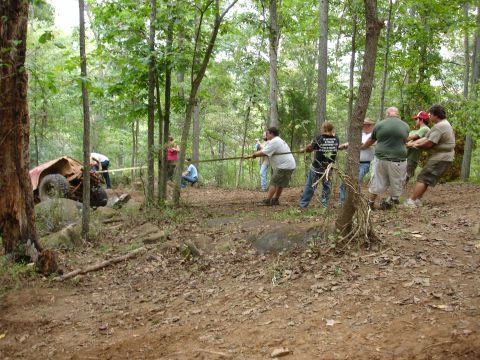 1996 Ford Bronco - A group of people attempt to pull a red off-road vehicle out of a wooded area where it appears to be stuck.