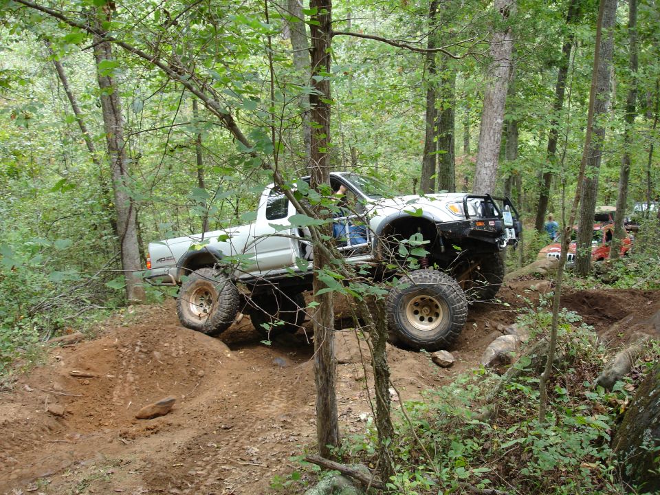 White pickup truck with large tires navigating an off-road trail in a forest