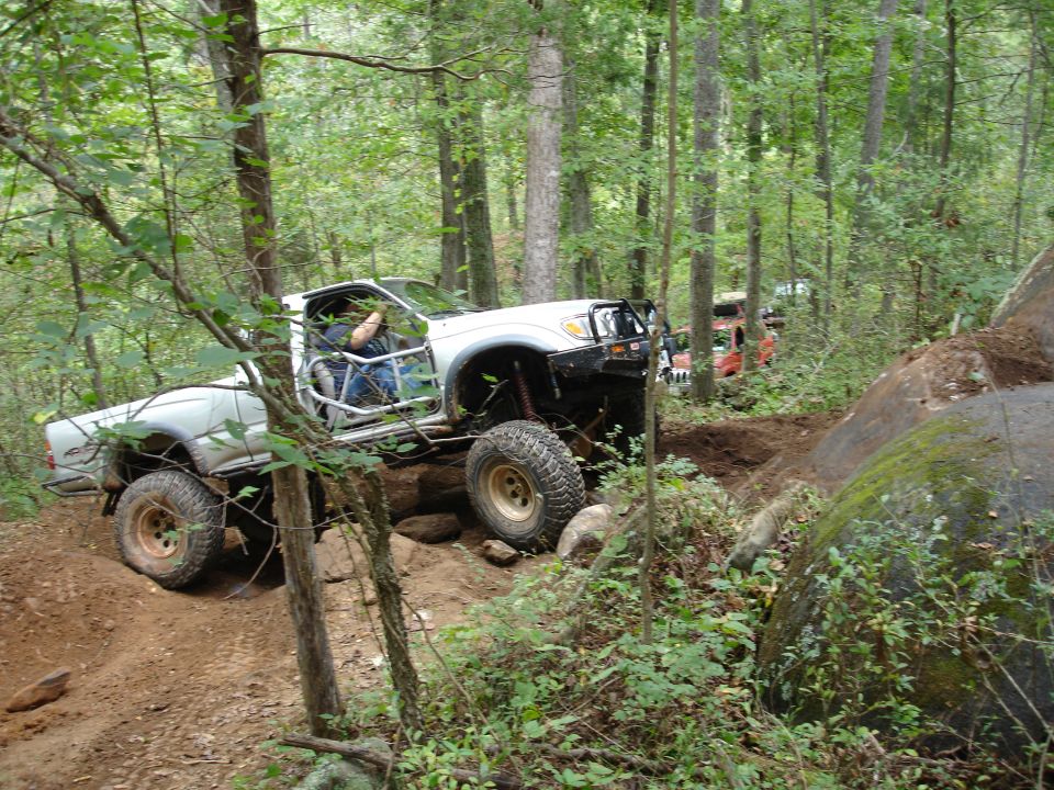 Off-road trucks on a dirt trail in a forest, one truck is white with a roll cage, another is orange