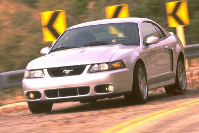 2003 Ford Mustang - A silver 2003 Ford Mustang is captured in motion on a winding road, with directional signs visible in the background.