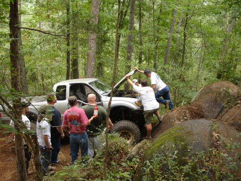 1996 Ford Bronco - Several people are gathered around a silver pickup truck, appearing to work on its engine while off-roading in a forest.