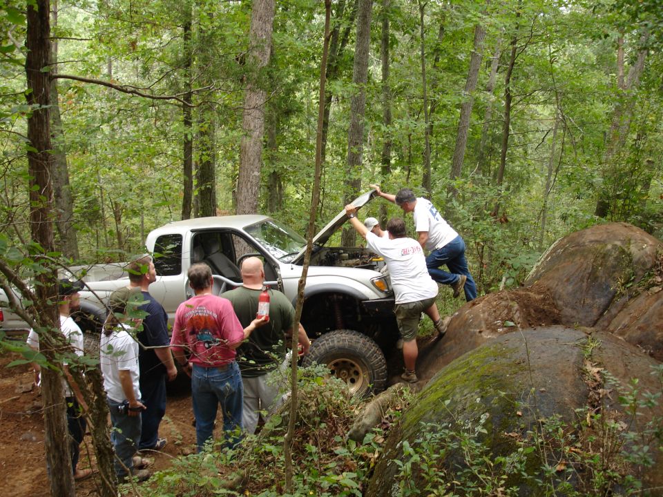 Group of guys gathered around a silver pickup truck with its hood open, working on the engine in a wooded area with large rocks