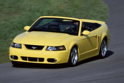 2003 Ford Mustang - A yellow 2003 Ford Mustang convertible is captured in motion on a paved road with a grassy embankment in the background.