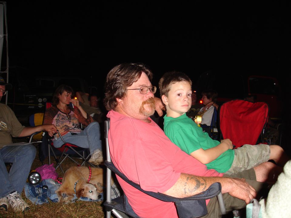 Man and boy sitting in chairs at night with other people in the background