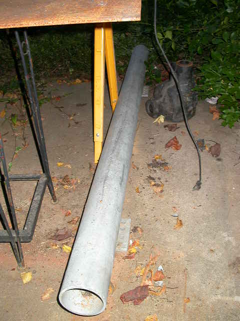 1990 Ford Bronco - A length of bare steel round tubing leaning against a rusty steel work table on a concrete pad, with a yellow sawhorse, a cast-iron part an…