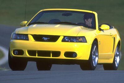 2003 Ford Mustang - A yellow 2003 Ford Mustang convertible is shown from the front, driving on a road with a driver visible.