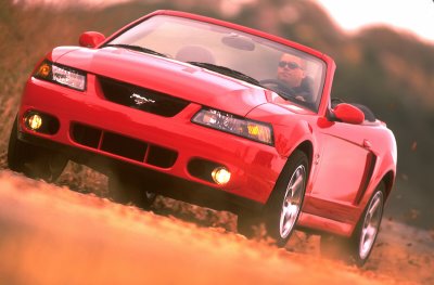 2003 Ford Mustang - A red 2003 Ford Mustang convertible is captured in a dynamic shot as it drives on a dirt road, kicking up dust.