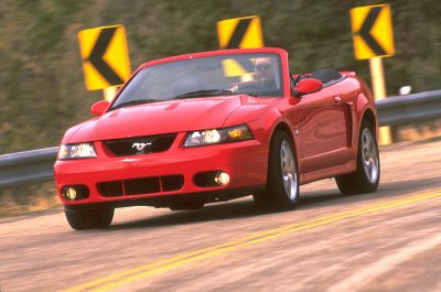 2003 Ford Mustang - Red 2003 Ford Mustang convertible driving on a road with yellow curve warning signs in the background.