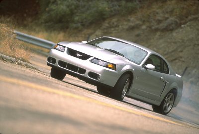 2003 Ford Mustang - A silver 2003 Ford Mustang is captured in motion on a winding road with a guardrail visible in the background.