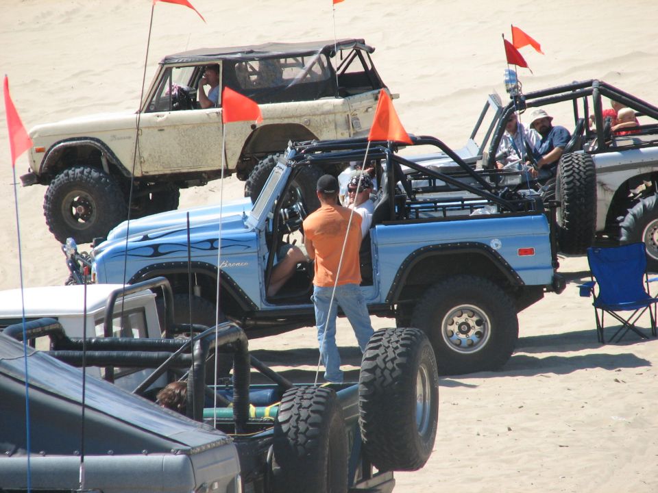 Light blue early Bronco being worked on among rigs with flags on the dunes