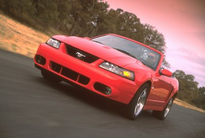 2003 Ford Mustang - A red 2003 Ford Mustang convertible drives on a road with trees in the background.