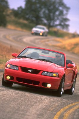 2003 Ford Mustang - A red 2003 Ford Mustang convertible cruises down a scenic, winding road with another vehicle in the distance.