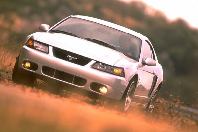 2003 Ford Mustang - A white 2003 Ford Mustang is captured in a dynamic shot, kicking up dust as it drives on a dirt road.