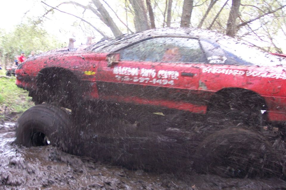 Red car stuck deep in mud, tires submerged. Text on door reads 'RACING PARTS CENTER 312-557-1494' and 'BASTARD BOILER'