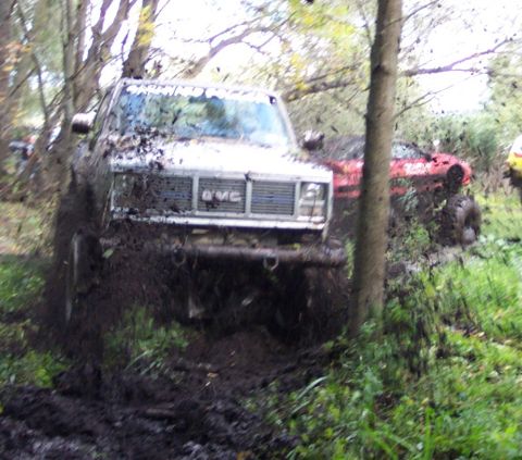 1978 Ford Bronco - A GMC truck is stuck in the mud, with another vehicle visible behind it in a wooded area.