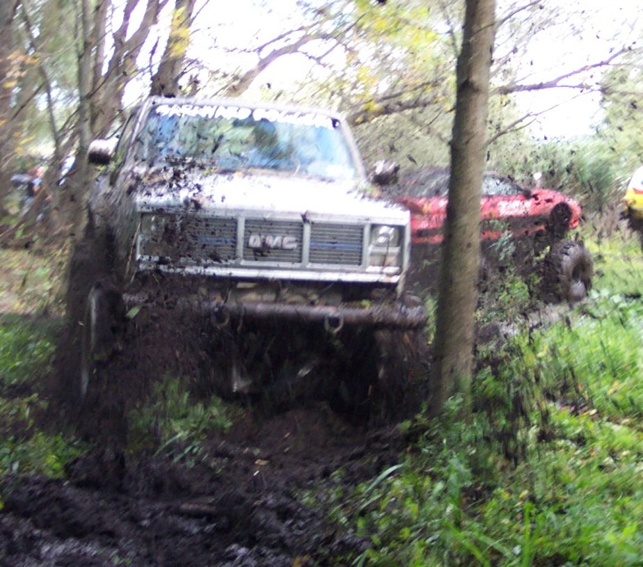 Muddy GMC truck stuck in woods, red vehicle behind it