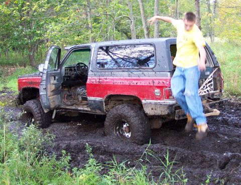 1978 Ford Bronco - A person jumps off the back of a red and gray SUV as it is stuck deep in the mud.