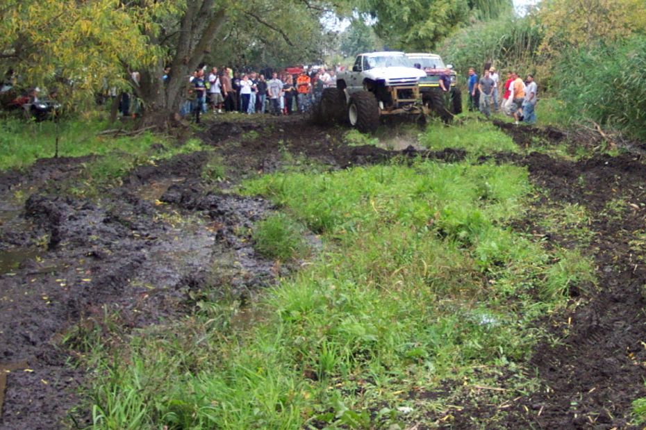Two modified trucks with large tires driving through a muddy field with spectators watching