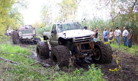 1978 Ford Bronco - Monster trucks bogged down in a muddy off-road course, watched by a group of onlookers.