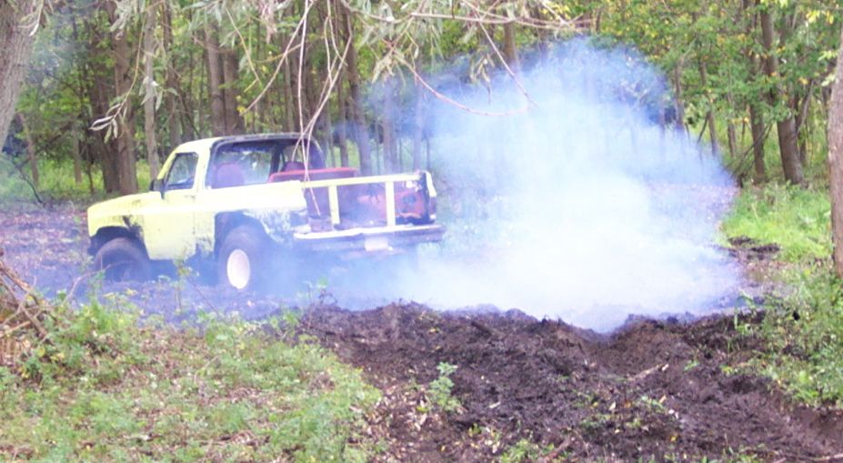 Yellow pickup truck with a white roof and bed rails, emitting white smoke while stuck in mud