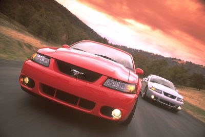 2003 Ford Mustang - Two Ford Mustangs drive on a road with a dramatic sunset sky.