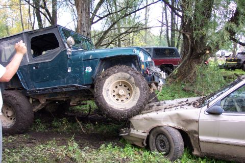 1978 Ford Bronco - A green Jeep is positioned on top of a damaged car, with other vehicles visible in the background, in a muddy, outdoor setting.