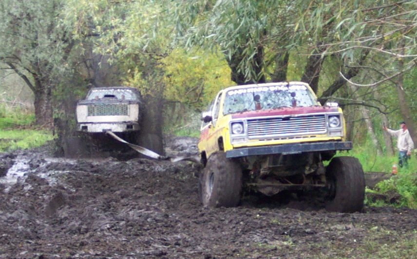 Yellow and red pickup truck with large tires in mud, front view. Another vehicle is stuck behind it
