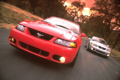 2003 Ford Mustang - A red 2003 Ford Mustang leads a silver Mustang on a winding road at sunset.