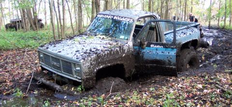 1978 Ford Bronco - A GMC pickup truck is deeply stuck in mud in a wooded area, with another vehicle visible in the background.